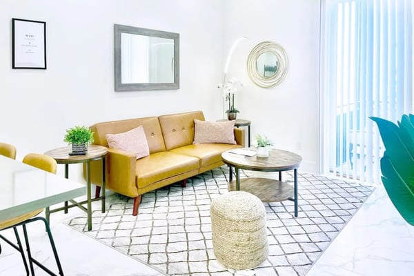 Bright living room with mustard yellow sofa, round coffee table, woven pouf, mirrors, and vertical blinds overlooking outdoor space