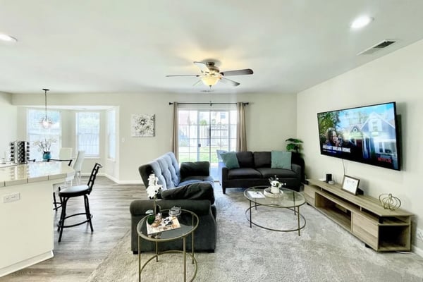 Open-plan living room with black sectional sofa, round glass coffee tables, wall-mounted TV, and glass doors overlooking backyard.